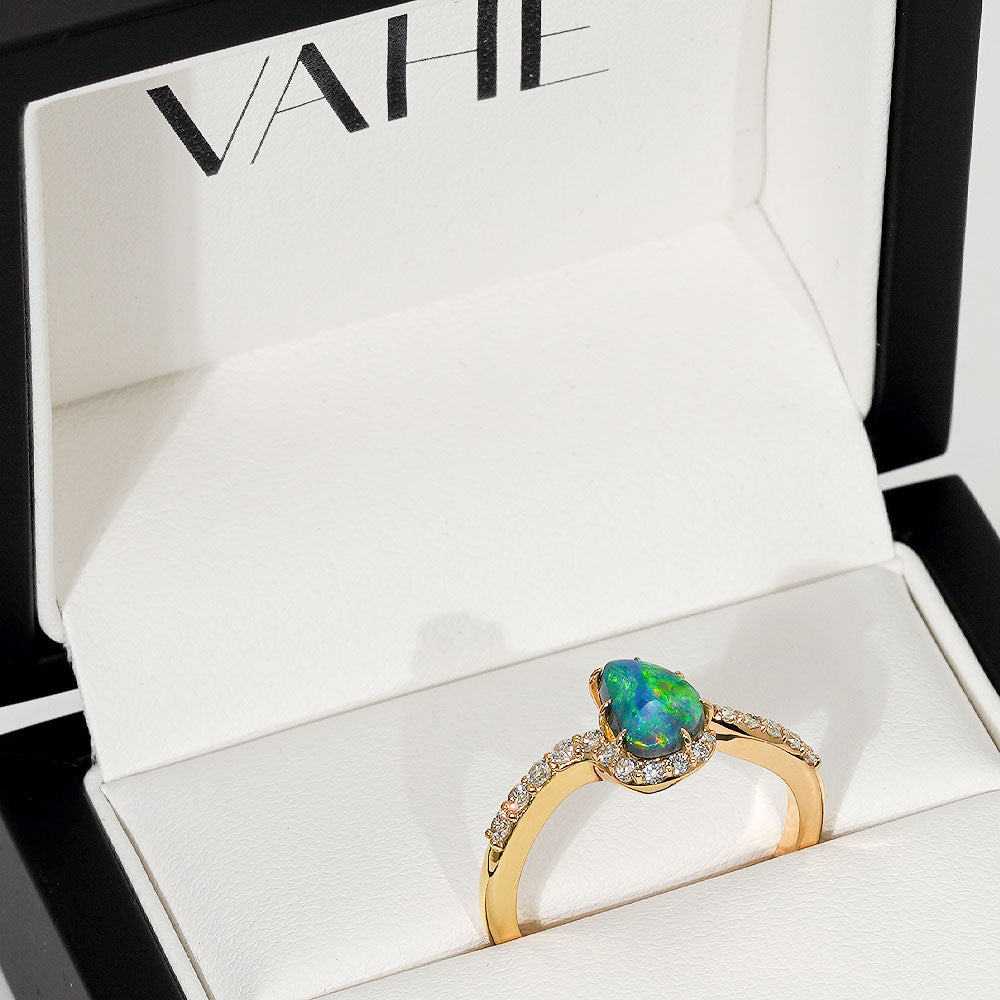 A gold ring with a teardrop-shaped iridescent opal and small clear stones, displayed inside an open white jewelry box with the brand name VAHE printed inside.