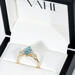 A gold ring with a pear-cut light blue gemstone and small clear accent stones, displayed inside an open white jewelry box with the brand name VAHE visible on the inner lid.