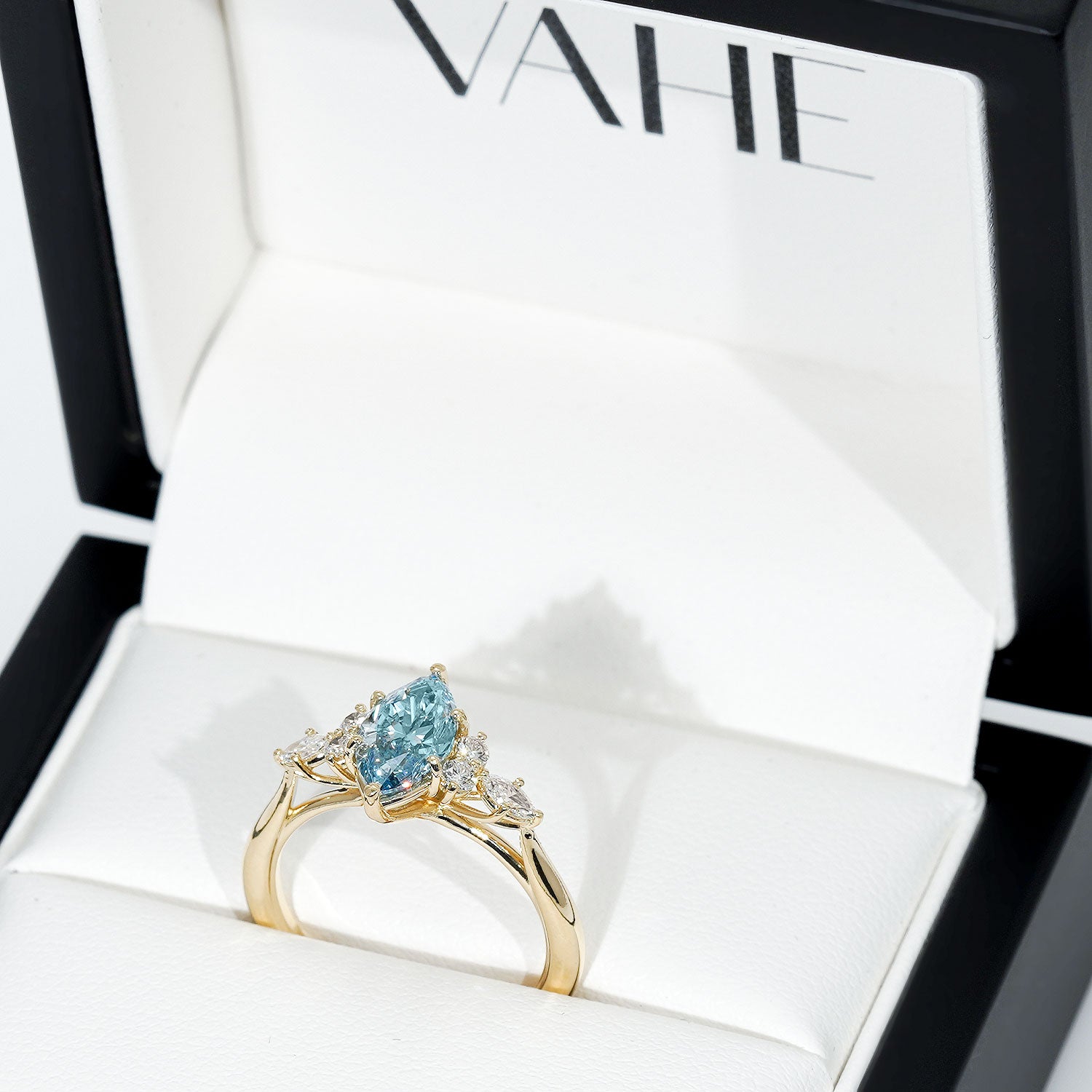 A gold ring with a pear-cut light blue gemstone and small clear accent stones, displayed inside an open white jewelry box with the brand name VAHE visible on the inner lid.