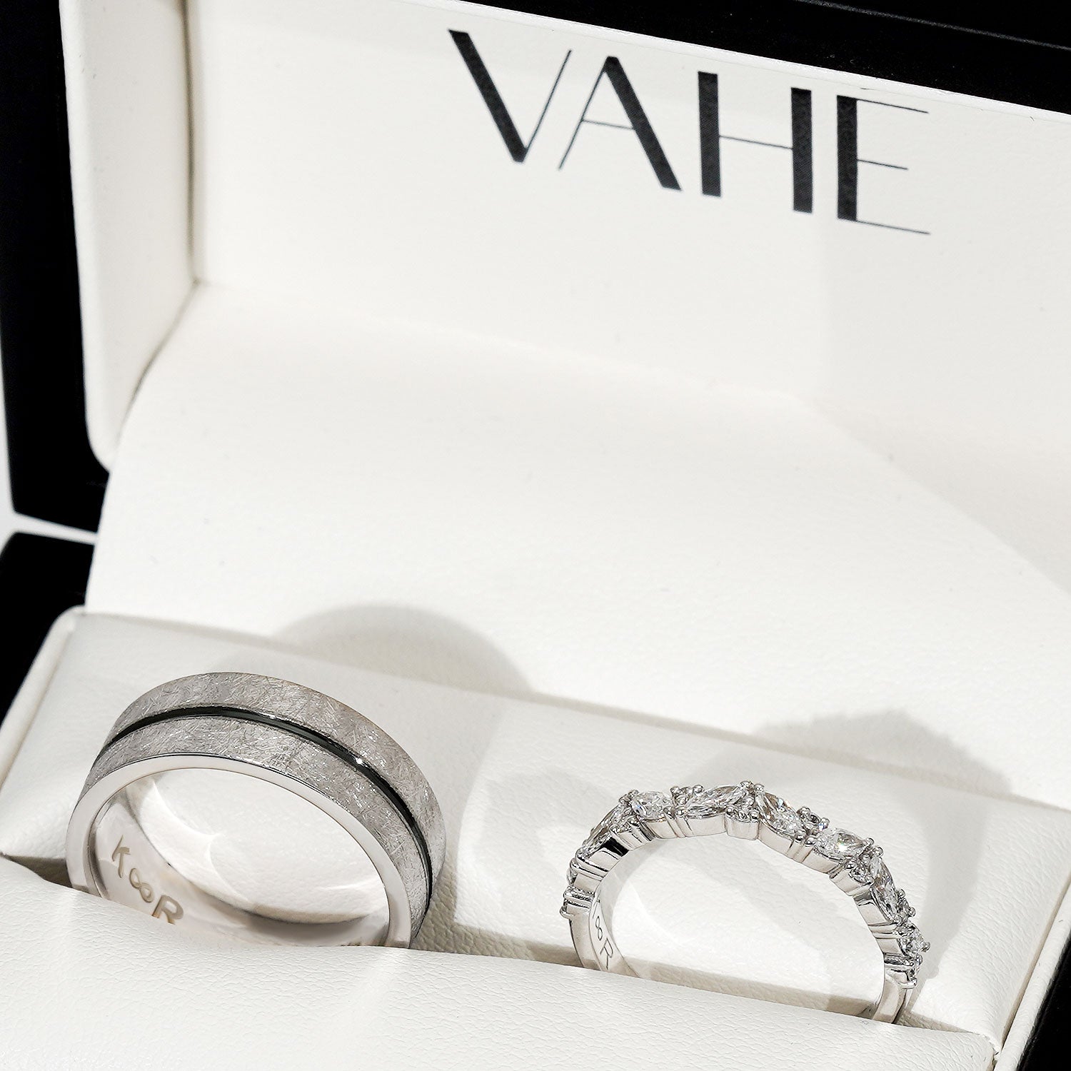 Two silver rings displayed inside an open white jewelry box with the brand name VAHE printed inside the lid. One ring has a textured band with a thin dark center stripe, and the other is a silver eternity-style ring set with clear stones.