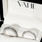 Two silver rings displayed inside an open white jewelry box with the brand name VAHE printed inside the lid. One ring has a textured band with a thin dark center stripe, and the other is a silver eternity-style ring set with clear stones.