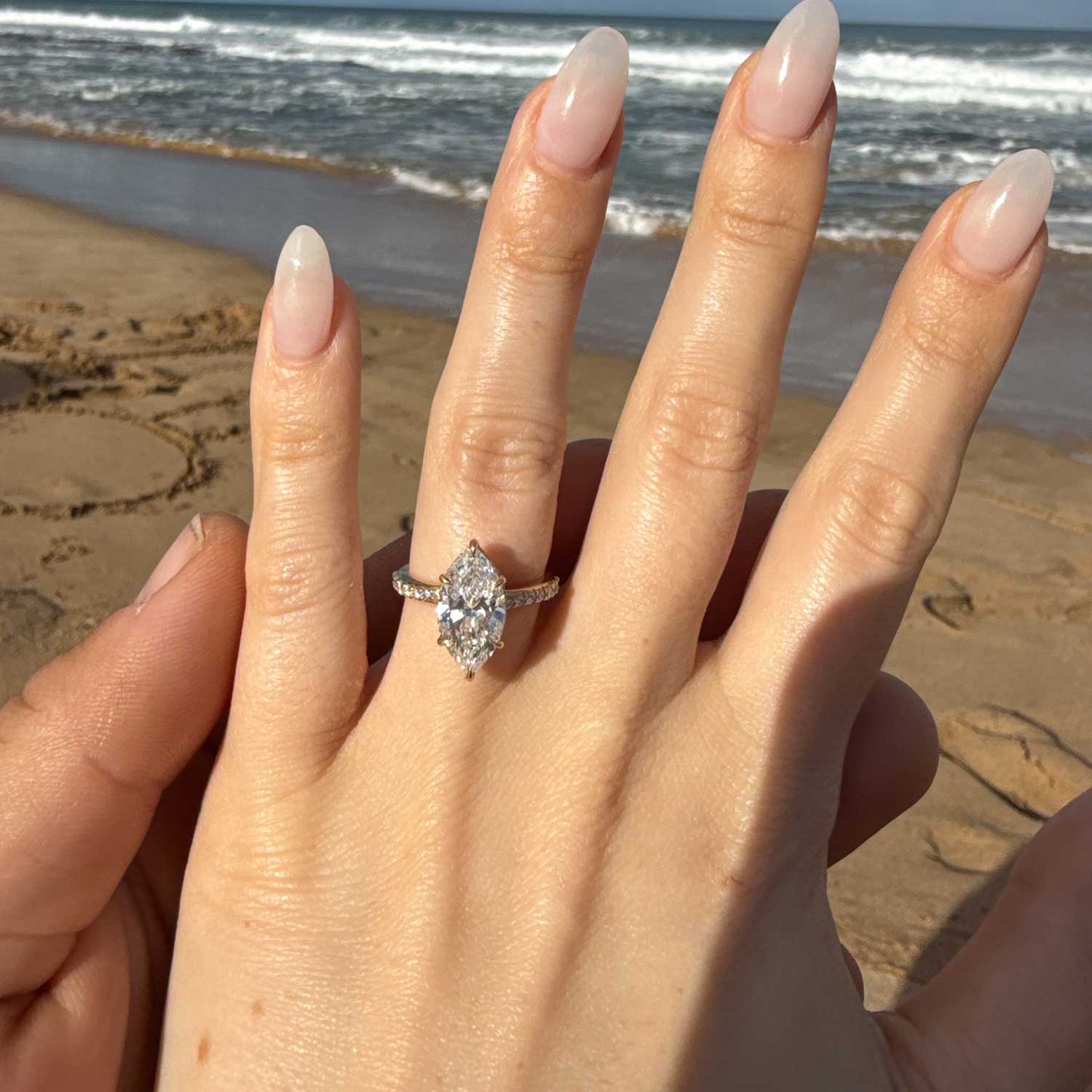 A silver-toned ring with a marquise-cut clear gemstone is shown on a person's hand, photographed outdoors on a sandy beach with the ocean in the background.