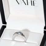A silver ring with a round light blue gemstone, displayed inside an open white jewelry box with the brand name VAHÉ visible on the inner lid.