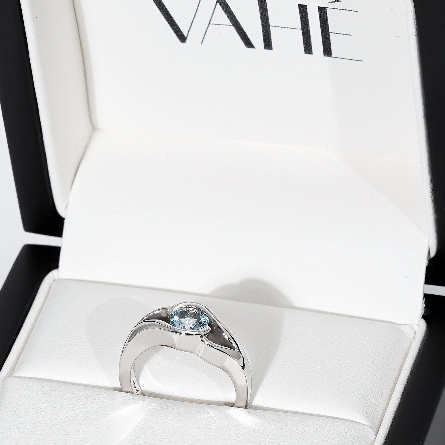 A silver ring with a round light blue gemstone, displayed inside an open white jewelry box with the brand name VAHÉ visible on the inner lid.