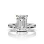 An emerald-cut diamond solitaire ring in a polished four-prong white gold or platinum setting, centered on a reflective white surface.
