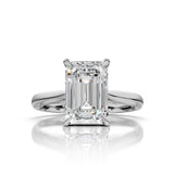An emerald-cut diamond solitaire ring in a polished four-prong white gold or platinum setting, centered on a reflective white surface.