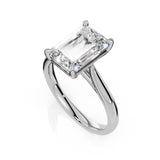 An emerald-cut diamond engagement ring in a polished silver-colored metal band, shown on a clean white background with a soft reflection.