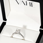 A silver engagement ring with a princess-cut clear gemstone displayed inside an open white jewelry box with the brand name VAHÉ printed on the inner lid.