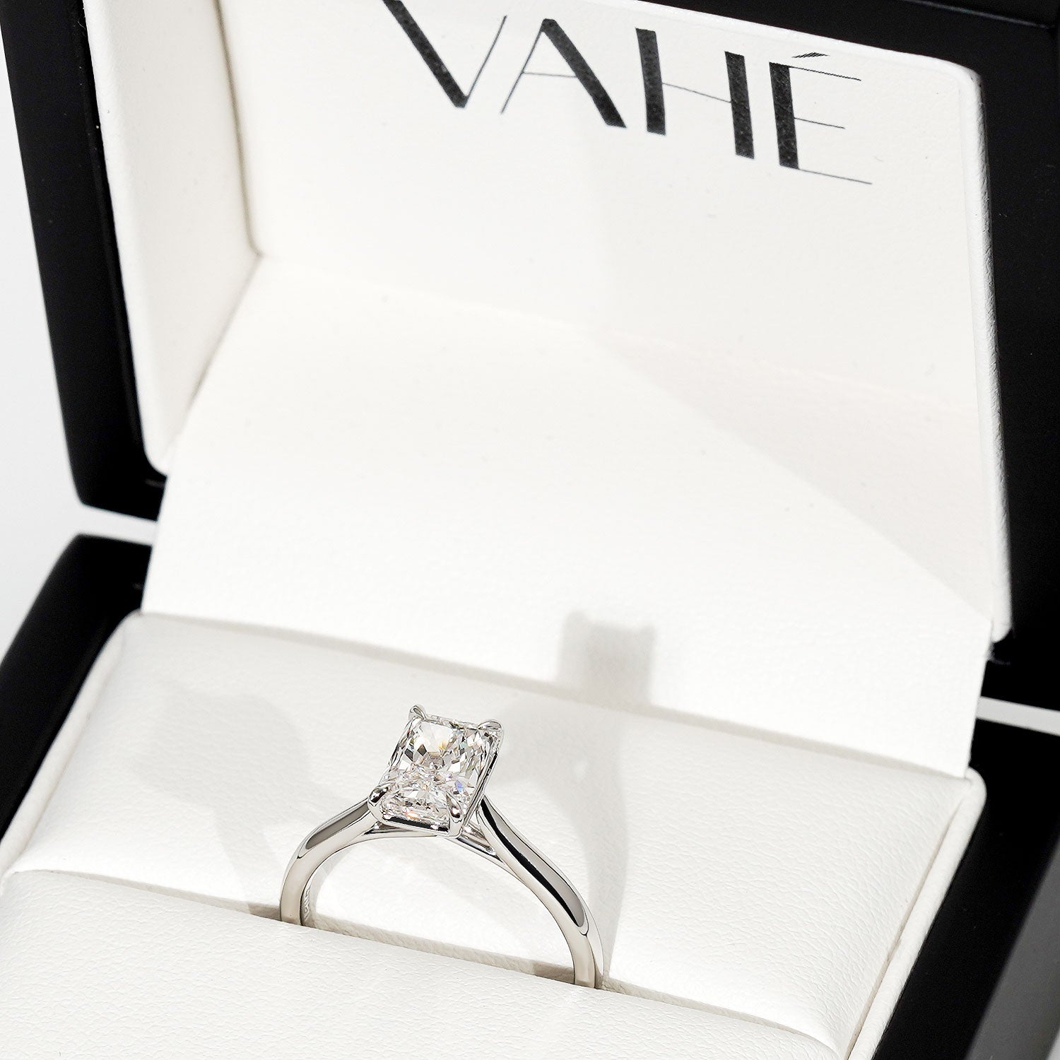 A silver engagement ring with a princess-cut clear gemstone displayed inside an open white jewelry box with the brand name VAHÉ printed on the inner lid.