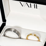 Two rings displayed inside a white jewelry box with the brand name VAHE printed on the inside lid. One ring is brushed silver-toned metal, and the other is yellow gold with a row of clear stones.