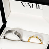 Two rings displayed inside a white jewelry box with the brand name VAHE printed on the inside lid. One ring is brushed silver-toned metal, and the other is yellow gold with a row of clear stones.