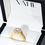 A yellow gold halo ring with a large round-cut center stone and smaller accent stones, displayed inside an open white jewelry box branded VAHE.