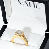A yellow gold halo ring with a large round-cut center stone and smaller accent stones, displayed inside an open white jewelry box branded VAHE.