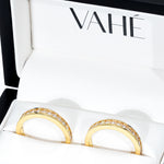 A pair of gold rings with a row of clear pavé stones set along the upper half, presented in a white jewelry box with the brand name VAHÉ visible inside.