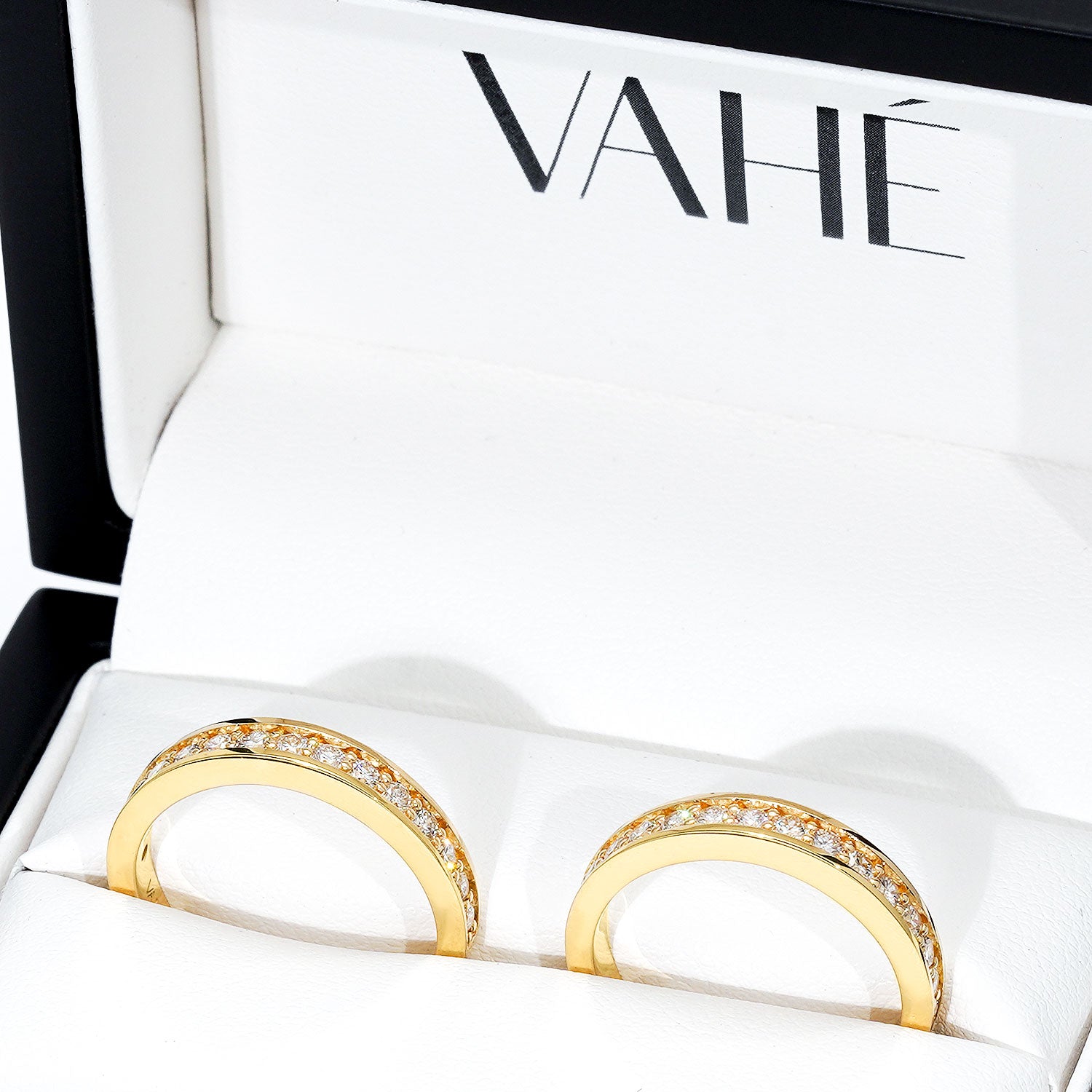A pair of gold rings with a row of clear pavé stones set along the upper half, presented in a white jewelry box with the brand name VAHÉ visible inside.