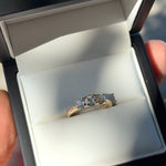 A three-stone diamond ring with light blue and clear stones set in a yellow gold band, displayed inside an open white jewelry box.