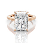 An emerald-cut diamond ring in a rose gold setting, displayed on a clean white reflective surface.