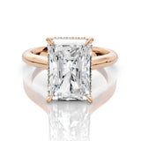 An emerald-cut diamond ring in a rose gold setting, displayed on a clean white reflective surface.
