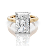 An emerald-cut diamond engagement ring with a yellow gold band and small diamond accents on the shoulders, set against a clean white background with a soft reflection.