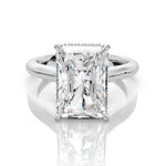 An emerald-cut diamond engagement ring in a silver-colored metal setting, shown on a reflective white surface.