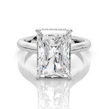An emerald-cut diamond engagement ring in a silver-colored metal setting, shown on a reflective white surface.