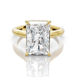 An emerald-cut diamond engagement ring with a yellow gold band and small diamond accents on the shoulders, shown on a reflective white surface.