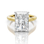 An emerald-cut diamond engagement ring with a yellow gold band and small diamond accents on the shoulders, set against a clean white background with a soft reflection.