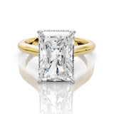 An emerald-cut diamond engagement ring with a yellow gold band and small diamond accents on the shoulders, set against a clean white background with a soft reflection.