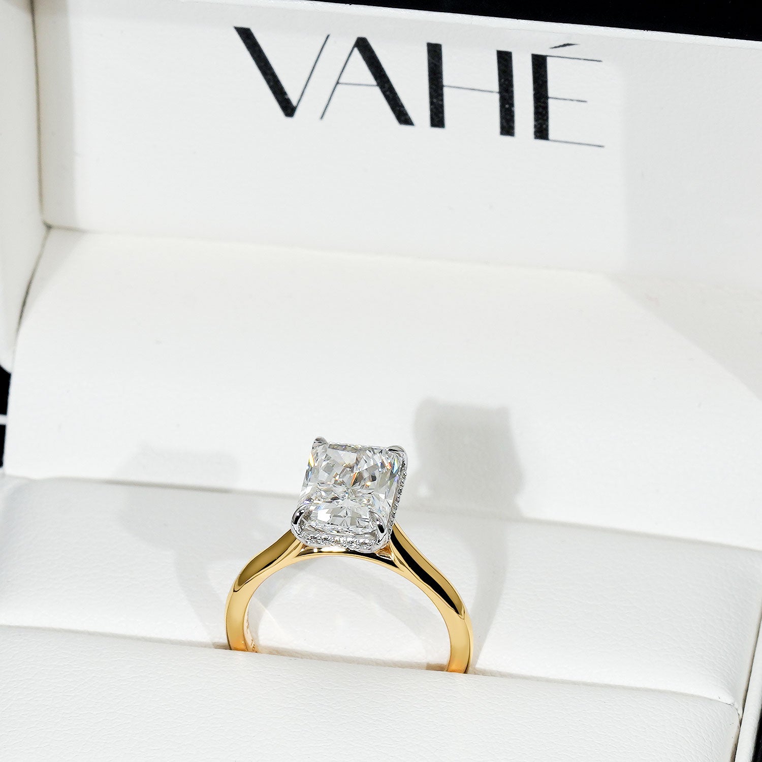A yellow gold ring with a square radiant-cut clear gemstone, displayed inside a white jewelry box with the brand name VAHÉ printed on the inside lid.