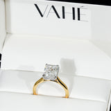 A yellow gold ring with a square radiant-cut clear gemstone, displayed inside a white jewelry box with the brand name VAHÉ printed on the inside lid.