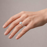 A close-up of a hand wearing a silver engagement ring with a round-cut diamond. The hand is shown against a solid, neutral taupe-gray background.
