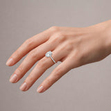 A close-up of a hand wearing a silver engagement ring with a round-cut diamond. The hand is shown against a solid, neutral taupe-gray background.