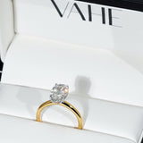 A gold ring with a round brilliant-cut clear gemstone in a prong setting, displayed inside a white jewelry box with the brand name VAHE printed on the inside lid.