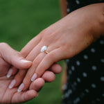 A close-up of hands holding, with a gold engagement ring featuring a round clear gemstone worn on one finger. The background is a soft, blurred green outdoor setting.