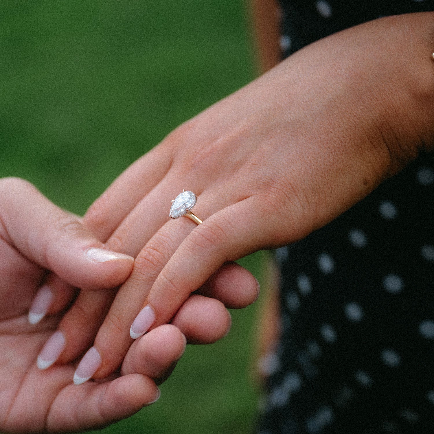 A close-up of hands holding, with a gold engagement ring featuring a round clear gemstone worn on one finger. The background is a soft, blurred green outdoor setting.